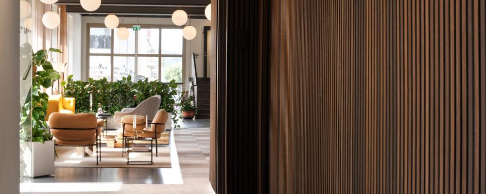 Träets markerade relief formar en taktil väggyta som markerar passagen och skapar en tydlig övergång. Väggen leder blick och rörelse vidare mot loungens mjuka sittgrupper och de runda pendellamporna, där ljus och möblering samverkar för att definiera rummets öppna karaktär.
Foto: Susanne E. Snaar