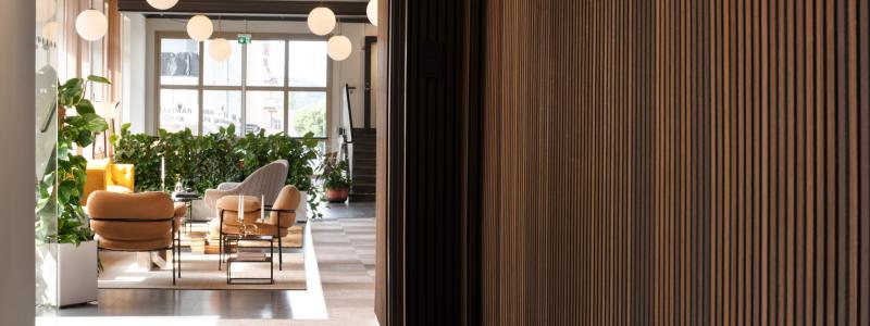 Träets markerade relief formar en taktil väggyta som markerar passagen och skapar en tydlig övergång. Väggen leder blick och rörelse vidare mot loungens mjuka sittgrupper och de runda pendellamporna, där ljus och möblering samverkar för att definiera rummets öppna karaktär.
Foto: Susanne E. Snaar