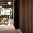 Träets markerade relief formar en taktil väggyta som markerar passagen och skapar en tydlig övergång. Väggen leder blick och rörelse vidare mot loungens mjuka sittgrupper och de runda pendellamporna, där ljus och möblering samverkar för att definiera rummets öppna karaktär.
Foto: Susanne E. Snaar