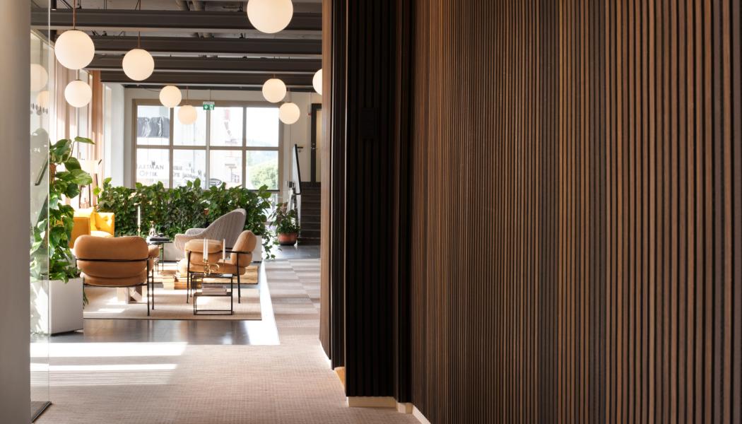 Träets markerade relief formar en taktil väggyta som markerar passagen och skapar en tydlig övergång. Väggen leder blick och rörelse vidare mot loungens mjuka sittgrupper och de runda pendellamporna, där ljus och möblering samverkar för att definiera rummets öppna karaktär.
Foto: Susanne E. Snaar
