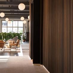 Träets markerade relief formar en taktil väggyta som markerar passagen och skapar en tydlig övergång. Väggen leder blick och rörelse vidare mot loungens mjuka sittgrupper och de runda pendellamporna, där ljus och möblering samverkar för att definiera rummets öppna karaktär.
Foto: Susanne E. Snaar